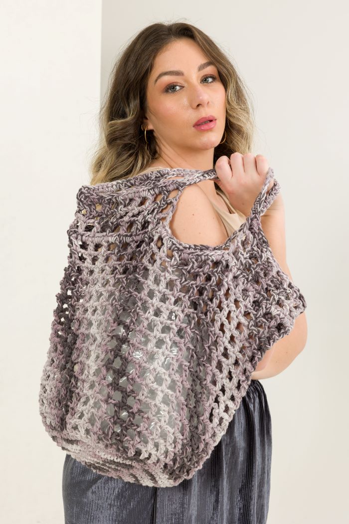 Círculo Natural Cotton Multicolor 4/6 – Cotton Yarn (226 m / 200 g) – A woman wearing a chunky, open-knit crocheted shoulder bag or tote in a heathered gray and taupe color, styled over a beige top and dark gray pants against a plain off-white background.