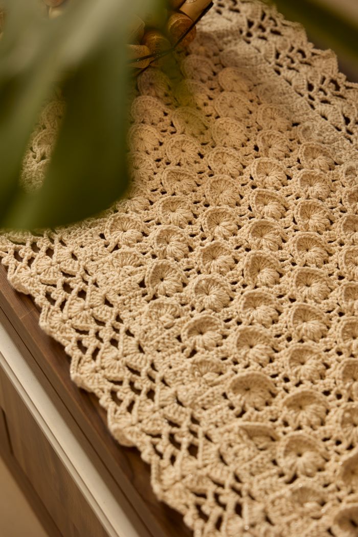 Circulo Barroco Natural Brilho - Natural Cotton Yarn Gold/Silver, 657m / 433m, 400g - A close-up of a beige crocheted lace table runner draped over a wooden surface, with soft green leaves partially framing the top left corner.