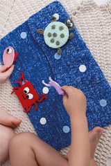 Círculo Amigurumi Pelúcia – Polyester Chenille Yarn (131 m / 85 g) - A close-up of a blue crocheted or knit fabric item with colorful sea-creature appliqués, including a green turtle, red crab, pink fish, and purple octopus, being held by a child's hands on a beige textured blanket.