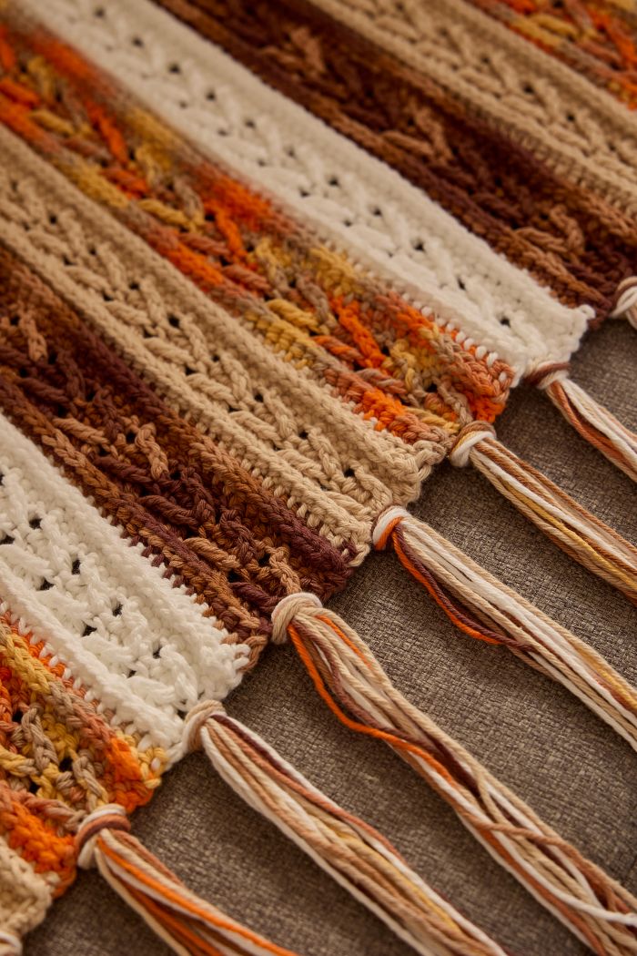 Círculo Natural Cotton Multicolor 4/6 – Cotton Yarn (226 m / 200 g) – A close-up of a crocheted or knitted textile with striped bands in cream, tan, brown, and orange, finished with long fringe tassels on a brown fabric background.