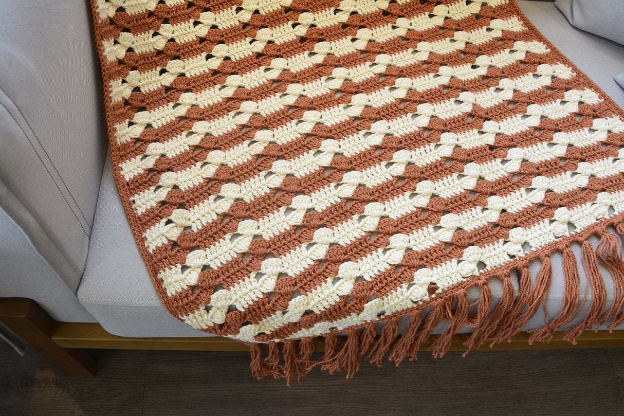Círculo Barroco Maxcolor Sparkle – Cotton Yarn (216m / 200g) - A close-up of a crocheted throw blanket draped over a light gray sofa. The blanket features a textured, raised chevron pattern in terracotta orange and cream, finished with a fringed edge.