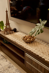 Circulo Barroco Natural Brilho  - Natural Cotton Yarn Gold/Silver, 657m / 433m, 400g - A cream-colored crocheted lace runner tablecloth draped over a dark wood TV stand in a living room setting, styled with a small Eiffel Tower figurine, a bowl of decorative stones, and a potted monstera plant.